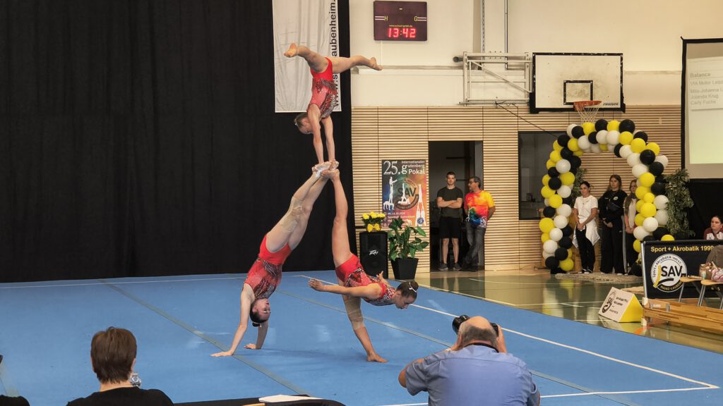 Zwei Sportlerinnen machen Handstand und halten sich quasi an den Füßen, auf denen die dritte einen Handstand mit gespreizten Beinen macht.